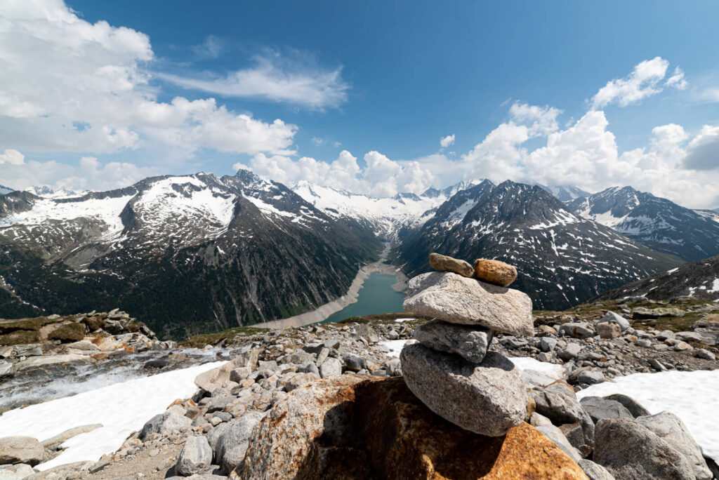 In der Tiefe liegt der Stausee, Berge im Hintergrund und im Vordergrund Steine. Es liegt in der Höhe Schnee